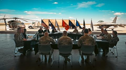 Rep. Crow sitting with military members with helicopters in the background